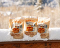 Three packages of Bonnie's Oatmeal Pancakes on a snowy surface with a blurred natural background