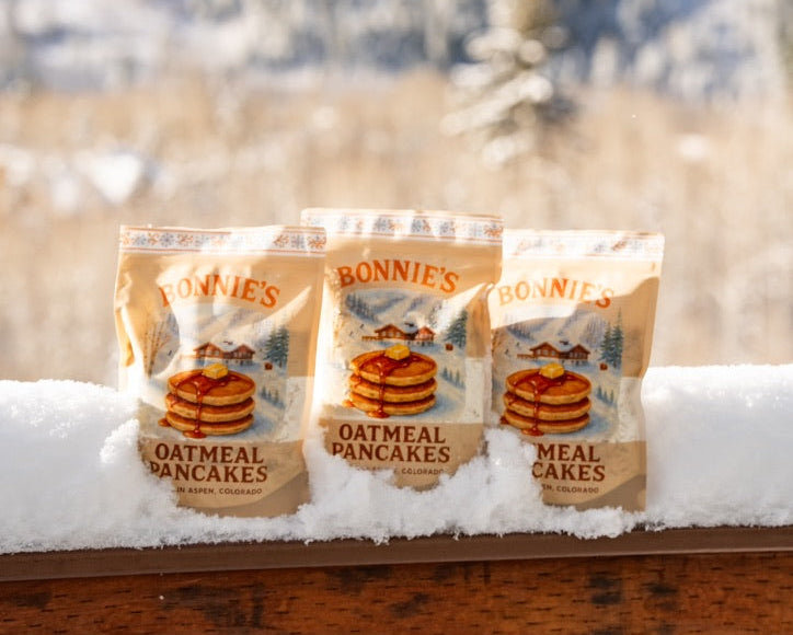 Three packages of Bonnie's Oatmeal Pancakes on a snowy surface with a blurred natural background