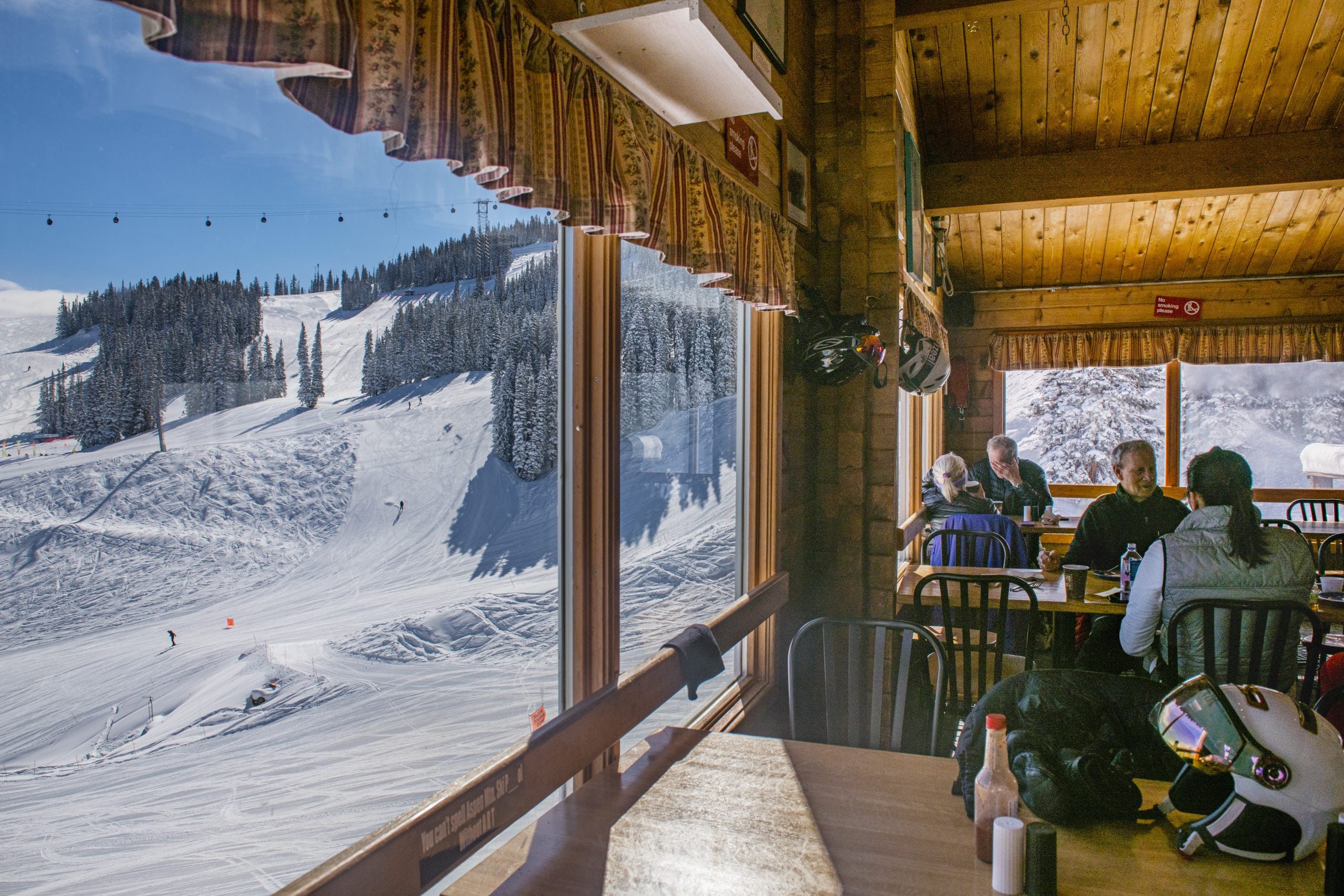 People sitting at a table inside a wooden cabin with a view of a snowy mountain landscape.