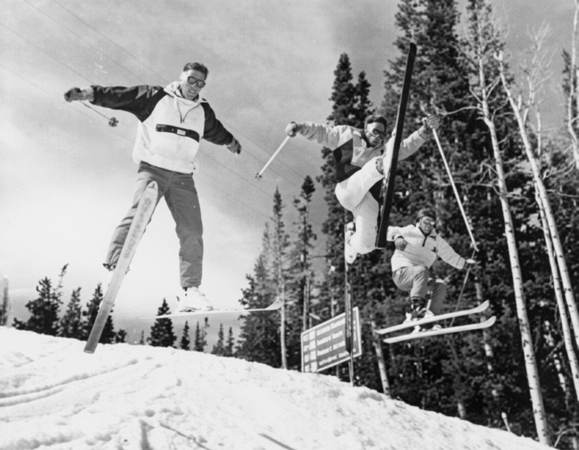Nick Malalet-Lee Clarke, Roland Topf, and Dominic Sanchez — three British ski bums who came to Aspen for the 1989–90 season to work and ski — catch air together on Aspen Mountain, 1990. Two of the three are on Dynastar skis. Behind them, a trail sign points toward Ruthie's Road, Ruthie's Restaurant, Bonnie's Restaurant, and the Silver Queen Gondola. The photo appears on page 327 of Mary Eshbaugh Hayes' The Story of Aspen, captioned "Triple airtime for Nick, Roland and Dominic."